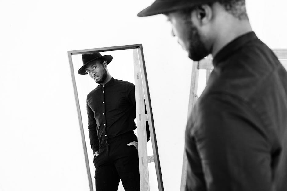 Black and white shot of reflection of thoughtful stylish African American male in shirt and hat looking in mirror on white background