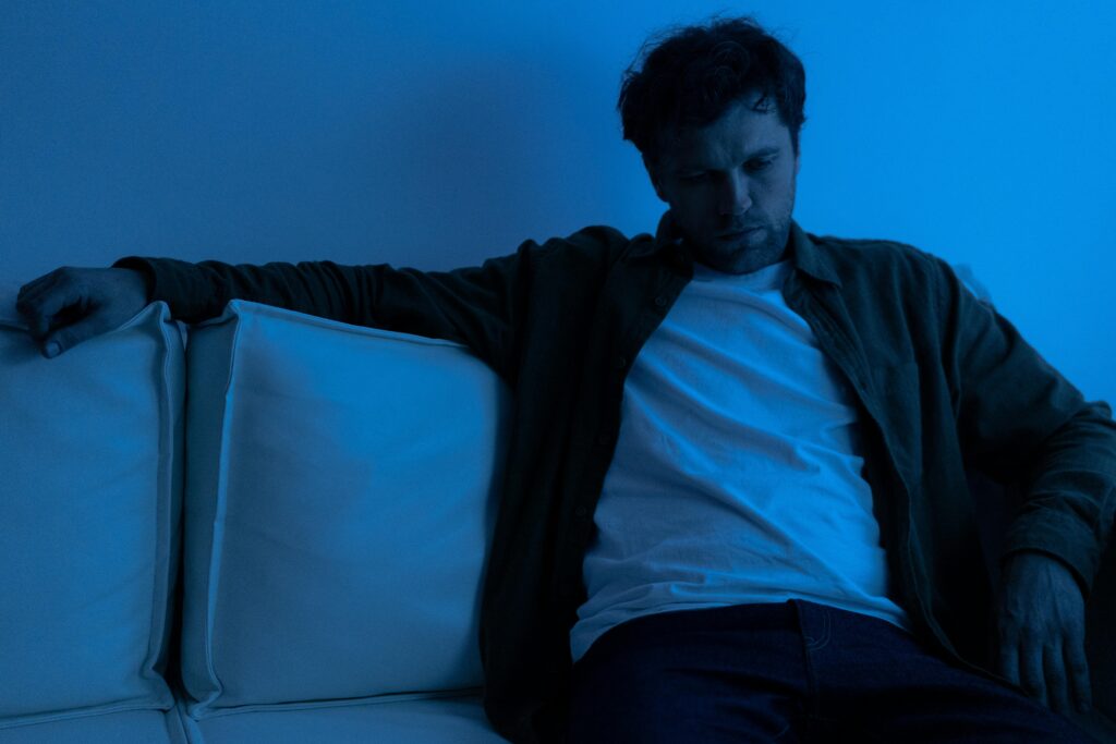 A man sitting alone on a sofa under blue lighting, conveying a somber mood.